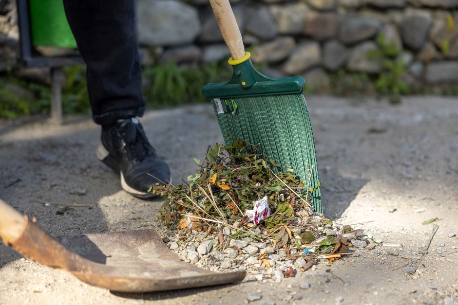 A person wearing black trousers and black sneakers is seen using a green hand broom with a wooden handle to sweep a pile of garden debris, including dried leaves, small twigs, and gravel, onto a dustpan on a paved outdoor surface. In the background, there is a stone wall with some green plants growing near it, and the area appears to be part of a garden or yard undergoing maintenance. The scene is lit with natural daylight, highlighting the textures of the soil, leaves, and paving stones. The image reflects garden maintenance activities that are relevant to the topic of garden waste rules for households, as discussed by Gardeners Hammersmith.