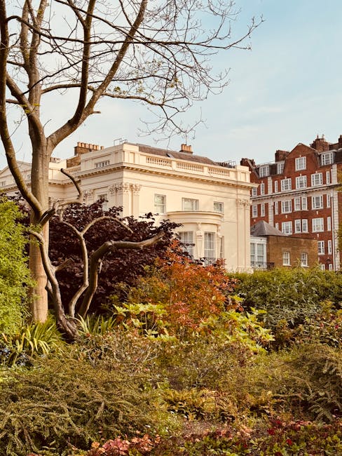 A well-maintained garden in front of a large, elegant white building with classical architectural details, including decorative cornices and large windows. The garden features a variety of plants, shrubs, and small trees, with some foliage showing autumnal colors like orange and deep red. A prominent leafless tree with twisting branches stands to the left, adding structure to the scene. The lawn is trimmed, and flowerbeds are filled with low-growing plants and ground cover. In the background, several brick residential buildings with white window frames and pitched roofs are visible. The lighting suggests a bright, partly cloudy day, highlighting the textures of the wet soil, leaves, and neatly maintained garden beds, reflecting typical garden maintenance and landscaping work conducted by Gardeners Hammersmith.