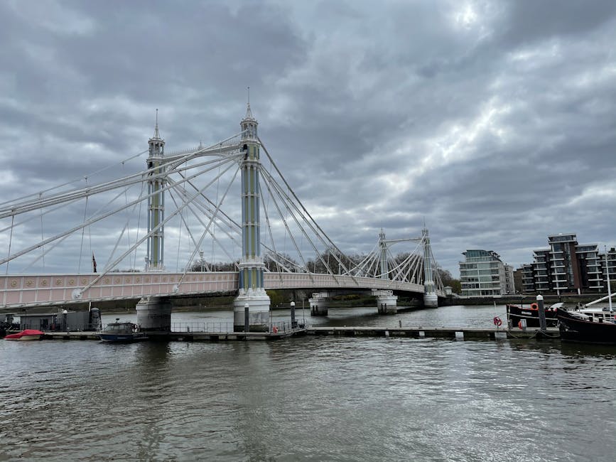 A view of the iconic Hammersmith Bridge spanning across the river, with its white-painted suspension cables and decorative towers visible beneath a cloudy sky. The water below appears slightly choppy, with boats docked along the riverbank on both sides. Modern residential buildings with glass facades line the opposite bank, creating a contrast with the historic bridge structure. The scene is set during daylight hours with diffuse lighting due to the overcast weather, and the overall atmosphere is calm, highlighting the bridge's architectural details and the surrounding urban waterscape. This image captures the bridge's prominent position as a landmark in Hammersmith, ideal for illustrating local outdoor scenery or regional landmarks.
